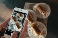 A person is holding a smartphone, capturing a photo of a tray with three tacos placed on it. The tacos are filled with ingredients that appear to include shredded vegetables and some kind of fried topping. The phone's screen displays the same tacos being photographed.