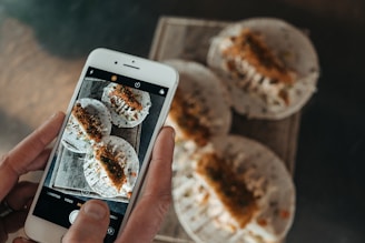 A person is holding a smartphone, capturing a photo of a tray with three tacos placed on it. The tacos are filled with ingredients that appear to include shredded vegetables and some kind of fried topping. The phone's screen displays the same tacos being photographed.