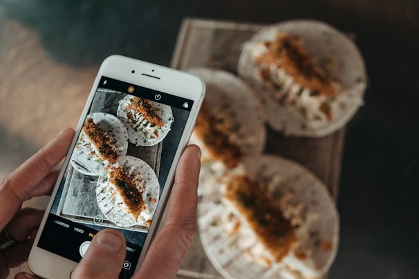 A person is holding a smartphone, capturing a photo of a tray with three tacos placed on it. The tacos are filled with ingredients that appear to include shredded vegetables and some kind of fried topping. The phone's screen displays the same tacos being photographed.