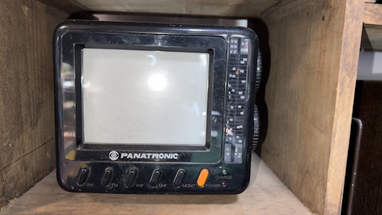 A small, vintage television set labeled 'PANATRONIC' placed inside a wooden shelf. The TV has a screen with surrounding buttons and dials for AM, FM, VHF, UHF, monitor, power, and charge functions.