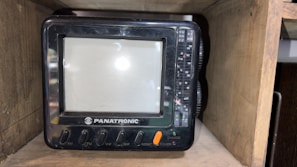 A small, vintage television set labeled 'PANATRONIC' placed inside a wooden shelf. The TV has a screen with surrounding buttons and dials for AM, FM, VHF, UHF, monitor, power, and charge functions.