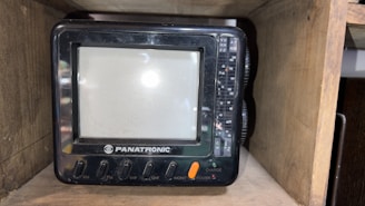 A small, vintage television set labeled 'PANATRONIC' placed inside a wooden shelf. The TV has a screen with surrounding buttons and dials for AM, FM, VHF, UHF, monitor, power, and charge functions.
