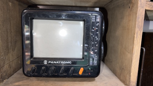 A small, vintage television set labeled 'PANATRONIC' placed inside a wooden shelf. The TV has a screen with surrounding buttons and dials for AM, FM, VHF, UHF, monitor, power, and charge functions.
