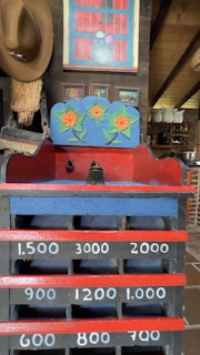 An old-fashioned wooden cash register with painted floral designs sits in an eclectic room. The register features hand-painted numerals and vibrant colors, including a decorative panel with orange flowers. A brown cowboy hat hangs on the wall, and paintings are displayed above. Shelves filled with vintage items are visible in the background.