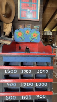 An old-fashioned wooden cash register with painted floral designs sits in an eclectic room. The register features hand-painted numerals and vibrant colors, including a decorative panel with orange flowers. A brown cowboy hat hangs on the wall, and paintings are displayed above. Shelves filled with vintage items are visible in the background.