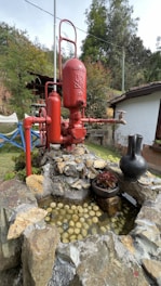 A large red industrial pump is positioned on a pile of rocks. To the right of the pump is a black jug placed on the rocky structure. In front of the pump, there is a shallow water feature filled with spherical objects that resemble small balls. The background includes a building with a white wall and a garden with various plants and trees.