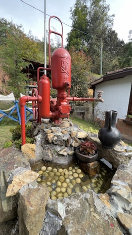 A large red industrial pump is positioned on a pile of rocks. To the right of the pump is a black jug placed on the rocky structure. In front of the pump, there is a shallow water feature filled with spherical objects that resemble small balls. The background includes a building with a white wall and a garden with various plants and trees.