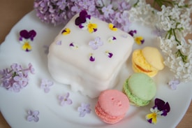 A square white cake adorned with delicate violets and other small purple flowers is placed on a white plate. Surrounding it are three colorful macarons in pastel shades of pink, green, and yellow. Nearby, clusters of lilac and white flowers add a natural touch to the arrangement.