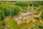 An aerial view of a large stone church with twin spires, surrounded by lush greenery and a cemetery in the foreground. The church features a metal roof and is set amidst a rural landscape with various houses and fields in the background.