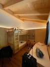 A cozy wooden room featuring a bunk bed with a person lying on the top bunk. The room is well-lit with warm lighting and has wooden walls and ceiling. There is a single bed with a pillow, two stools, and a hanging lamp. On the floor, there's a suitcase and a backpack placed near the bed.