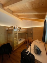 A cozy wooden room featuring a bunk bed with a person lying on the top bunk. The room is well-lit with warm lighting and has wooden walls and ceiling. There is a single bed with a pillow, two stools, and a hanging lamp. On the floor, there's a suitcase and a backpack placed near the bed.