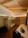 A cozy wooden room featuring a bunk bed with a person lying on the top bunk. The room is well-lit with warm lighting and has wooden walls and ceiling. There is a single bed with a pillow, two stools, and a hanging lamp. On the floor, there's a suitcase and a backpack placed near the bed.