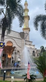 A tall minaret next to a detailed stone building with arches. A fountain with water jets is positioned in front, surrounded by Malaysian flags and palm trees. The cloudy sky forms the backdrop.