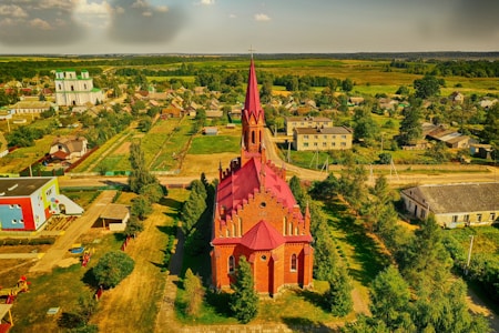 An aerial view showcases a vibrant, red-bricked church with a tall spire surrounded by lush greenery and a small village. The architecture stands out prominently amidst houses and open fields extending toward the horizon. A colorful playground and modern buildings nearby add a contrast to the traditional rural setting.