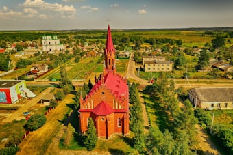 An aerial view of a countryside village featuring a prominent red-roofed brick church with a tall spire, surrounded by greenery and other village buildings. The landscape includes scattered houses, trees, and fields extending to the horizon.
