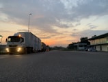 A semi-truck is parked on a road during early morning or evening, with a colorful sky featuring hues of orange and blue. The scene includes a few street lamps, buildings on the right, and some greenery.