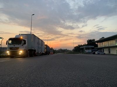 A semi-truck is parked on a road during early morning or evening, with a colorful sky featuring hues of orange and blue. The scene includes a few street lamps, buildings on the right, and some greenery.