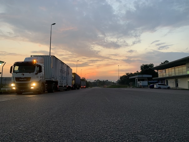 A semi-truck is parked on a road during early morning or evening, with a colorful sky featuring hues of orange and blue. The scene includes a few street lamps, buildings on the right, and some greenery.