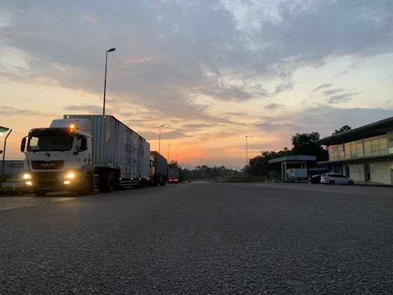 A semi-truck is parked on a road during early morning or evening, with a colorful sky featuring hues of orange and blue. The scene includes a few street lamps, buildings on the right, and some greenery.