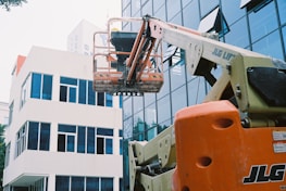 A construction lift with an orange base extends towards the upper levels of a modern glass building. The lift's platform is positioned in front of the reflective windows, and a white adjacent building is visible. The scene suggests maintenance or construction work in an urban setting.