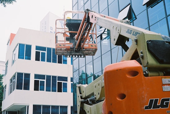 A construction lift with an orange base extends towards the upper levels of a modern glass building. The lift's platform is positioned in front of the reflective windows, and a white adjacent building is visible. The scene suggests maintenance or construction work in an urban setting.