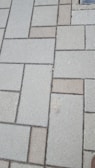 Concrete paving stones arranged in a rectangular pattern, with a mix of light and dark shades. The surface appears slightly rough and weathered.