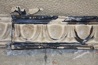 A section of an ornate, textured molding displaying signs of age and deterioration. The surface appears cracked and chipped, revealing underlying darker layers beneath the original beige or tan finish. The design features repeating round shapes adorned with dark, irregular patterns and edges.