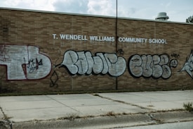 A brick wall of a community school is covered with large, stylized graffiti featuring bubble letters in black and silver. The school name, T. Wendell Williams Community School, is displayed in white block letters above the graffiti. The graffiti includes various tags and symbols.