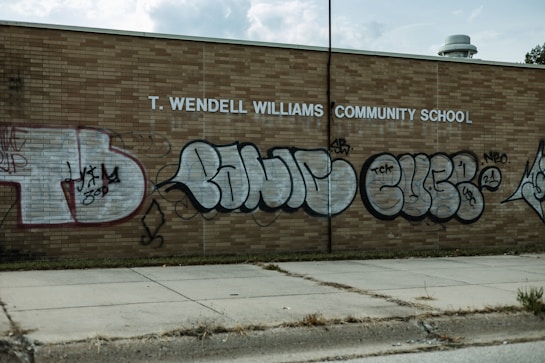 A brick wall of a community school is covered with large, stylized graffiti featuring bubble letters in black and silver. The school name, T. Wendell Williams Community School, is displayed in white block letters above the graffiti. The graffiti includes various tags and symbols.