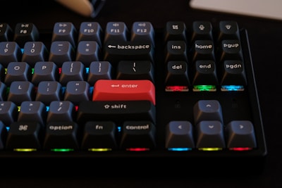 A mechanical keyboard featuring dark keycaps with white lettering and several keys illuminated by RGB lighting in different colors. The enter key stands out in bright red, surrounded by other standard keys like control, shift, and option. The overall color scheme is dark with colorful accents.