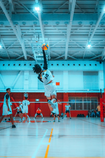 A volleyball player is leaping mid-air to hit a volleyball in an indoor court. The focus is on the athlete's athleticism and dynamic movement. There are other players in the background, indicating a game setting.