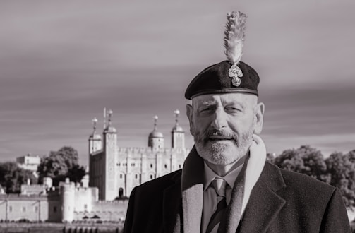 A man wearing a suit, coat, and a beret with a feather stands in front of a historic castle under a cloudy sky. The image is in black and white, highlighting the architectural details of the castle and the man's formal attire.