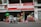 In front of a small shop, various colorful plastic items, including chairs, bins, and containers, are displayed for sale. A person rides a bicycle with large bags attached, passing by the shop on the street.