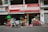 In front of a small shop, various colorful plastic items, including chairs, bins, and containers, are displayed for sale. A person rides a bicycle with large bags attached, passing by the shop on the street.