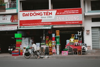 In front of a small shop, various colorful plastic items, including chairs, bins, and containers, are displayed for sale. A person rides a bicycle with large bags attached, passing by the shop on the street.
