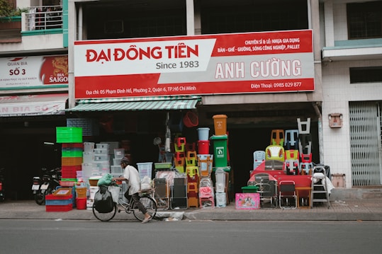 In front of a small shop, various colorful plastic items, including chairs, bins, and containers, are displayed for sale. A person rides a bicycle with large bags attached, passing by the shop on the street.