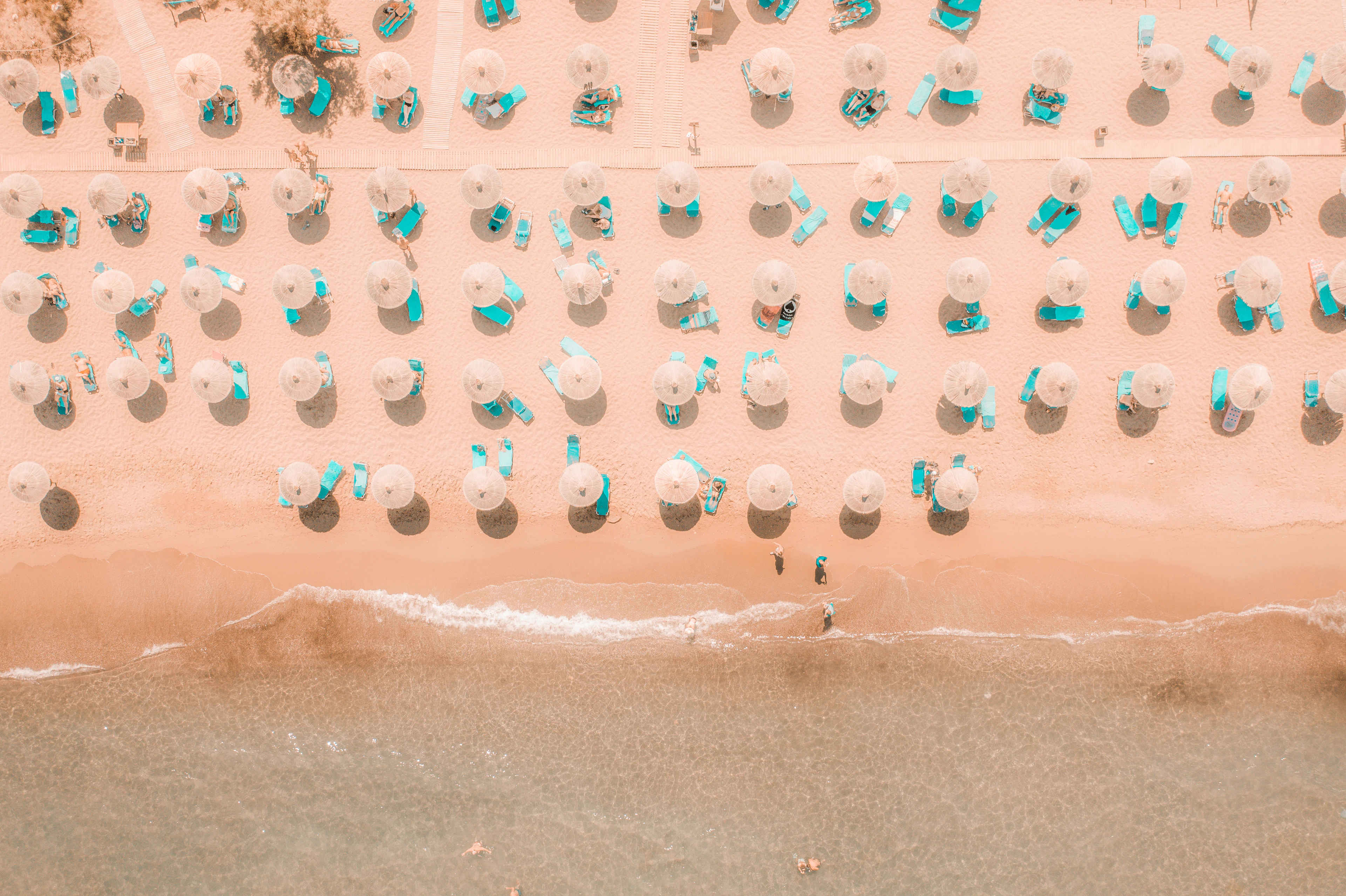 Rows of sun loungers and umbrellas line a sandy Cretan beach beside turquoise shallows, seen from above.