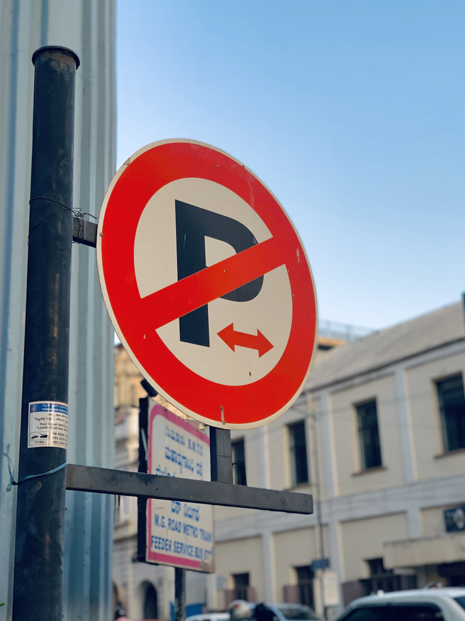 The image shows a red and white no parking sign mounted on a black pole, affixed to a building with a blue sky backdrop. The sign is clearly visible against the urban landscape, indicating that parking is not allowed in the area. In the background, there are several cars parked along the street, suggesting that this location may be a busy or popular spot for drivers.