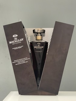 A luxurious bottle of Macallan Reflexion whisky is elegantly displayed in an open black box. The bottle has a sleek, dark design with a golden cap, and the box is designed with subtle, intricate patterns. The packaging exudes sophistication and exclusivity.