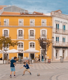 Two children are playing with a soccer ball in a patterned plaza in front of a bright yellow building with white-trimmed windows. The scene includes several people walking and sitting near a café with umbrellas on a sunny day.