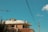 A building under construction with a worker on the roof. The sky is clear with minimal clouds, and a tall crane is visible on the left side. Power lines cross the image diagonally, and a streetlight is visible on the right.