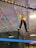 A person wearing bright athletic clothing navigates a suspended obstacle course indoors, balancing on ropes and utilizing a harness for safety. The background features industrial ceiling panels and lighting, adding to the adventurous atmosphere.