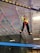 A person wearing bright athletic clothing navigates a suspended obstacle course indoors, balancing on ropes and utilizing a harness for safety. The background features industrial ceiling panels and lighting, adding to the adventurous atmosphere.