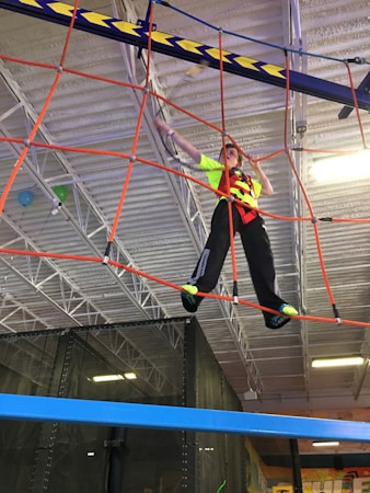 A person wearing bright athletic clothing navigates a suspended obstacle course indoors, balancing on ropes and utilizing a harness for safety. The background features industrial ceiling panels and lighting, adding to the adventurous atmosphere.