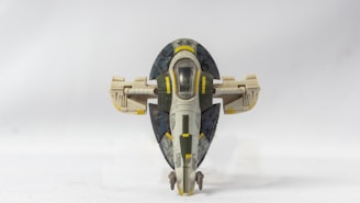 A detailed model of a spacecraft with a curved, oval-shaped design. It features a central cockpit with transparent windows and distinct yellow, green, and beige markings. The background is a plain white surface, highlighting the model.