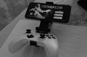 A mobile phone is mounted on a stand displaying a game titled 'Grimvalor' with options such as 'Continue', 'New Game', and 'Extras'. In front of the phone, there is a game controller placed on a table. The setup indicates gaming activity possibly using a smartphone as the screen.