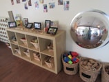 A wooden shelf unit contains wicker baskets filled with toys and materials, with framed photographs displayed on top. A variety of children's artwork and photos are attached to the wall above the shelf. To the right of the shelf, there is a large convex mirror reflecting the room, and on the floor, several baskets are filled with colorful building blocks and wooden pieces.