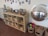 A wooden shelf unit contains wicker baskets filled with toys and materials, with framed photographs displayed on top. A variety of children's artwork and photos are attached to the wall above the shelf. To the right of the shelf, there is a large convex mirror reflecting the room, and on the floor, several baskets are filled with colorful building blocks and wooden pieces.