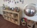 A wooden shelf unit contains wicker baskets filled with toys and materials, with framed photographs displayed on top. A variety of children's artwork and photos are attached to the wall above the shelf. To the right of the shelf, there is a large convex mirror reflecting the room, and on the floor, several baskets are filled with colorful building blocks and wooden pieces.