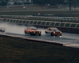 Dramatic wide shot of F1 cars racing closely on a wet circuit at sunset with dark clouds overhead.